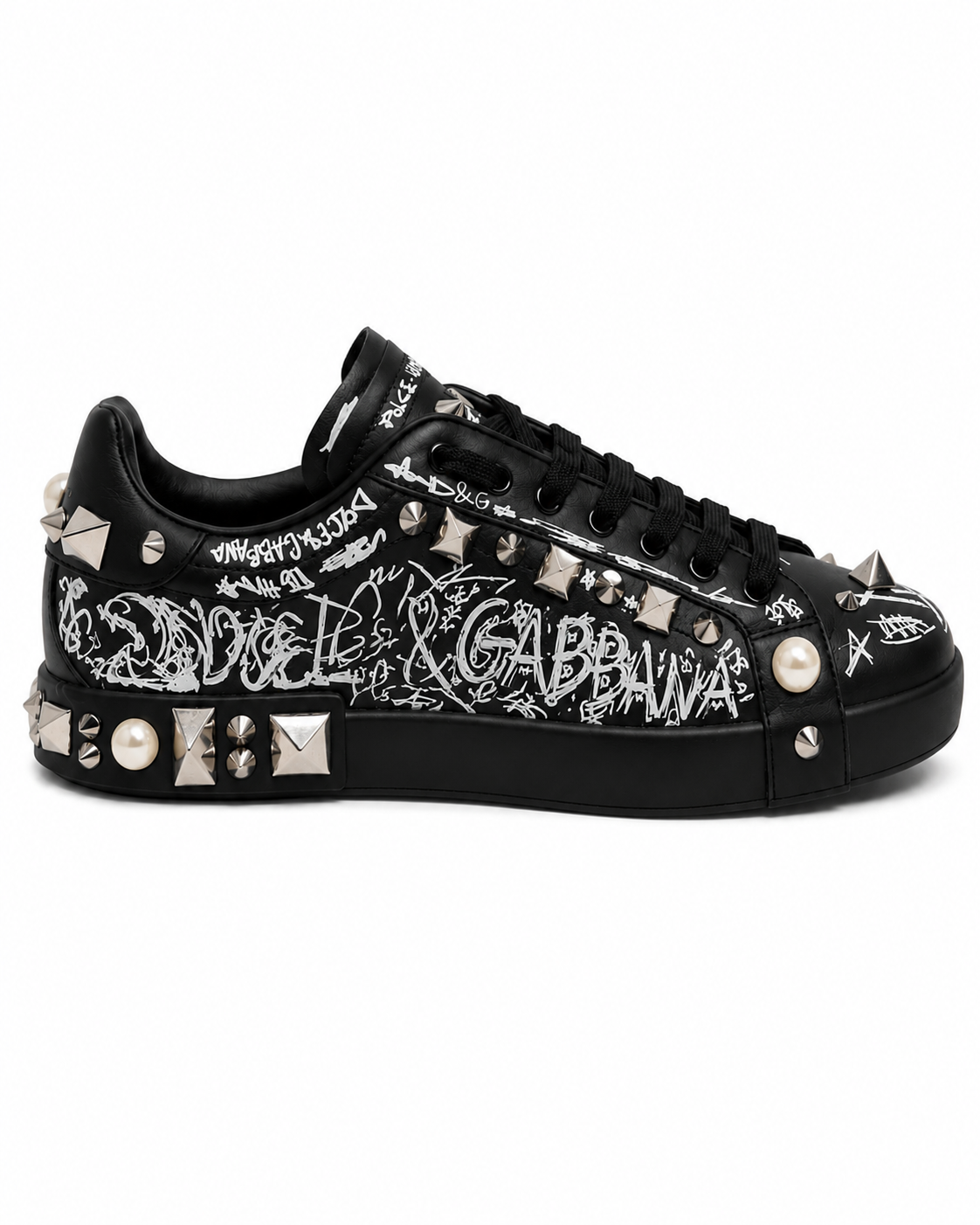 A black low-top sneaker by Dolce & Gabbana featuring white graffiti-style script graffiti across the side, silver metal studs, spikes, and a white faux-pearl accent on the toe box, photographed against a plain white background.