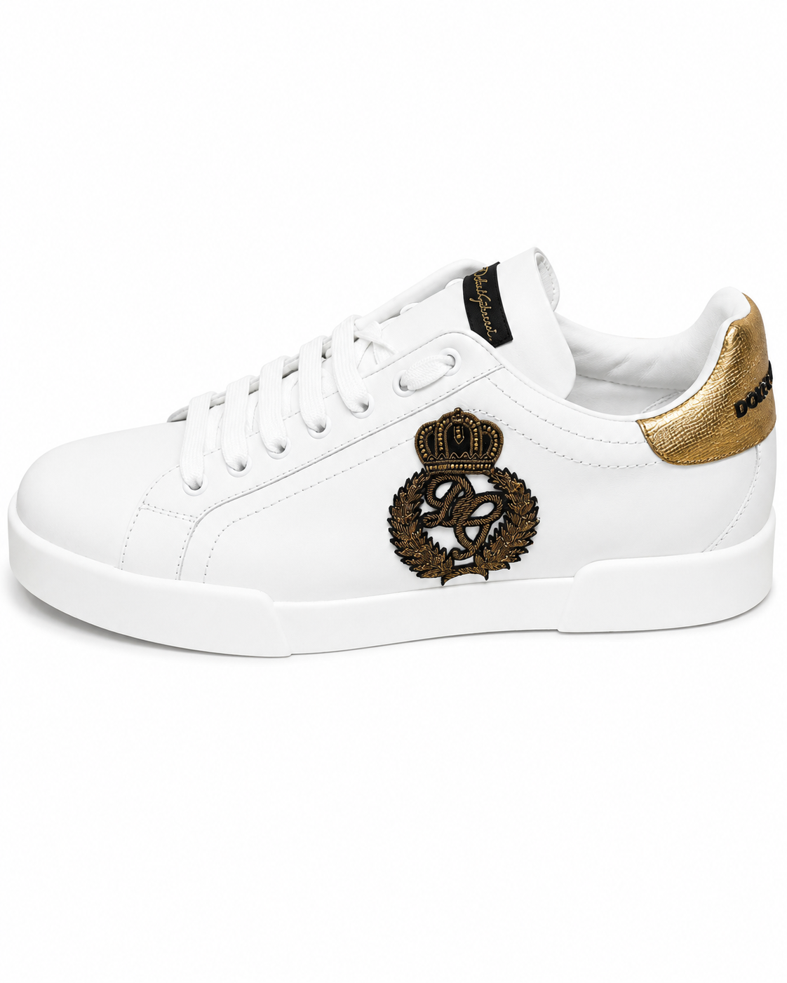 A white leather low-top sneaker with a metallic gold heel tab, white laces, and a prominent black and gold embroidered crest on the side.