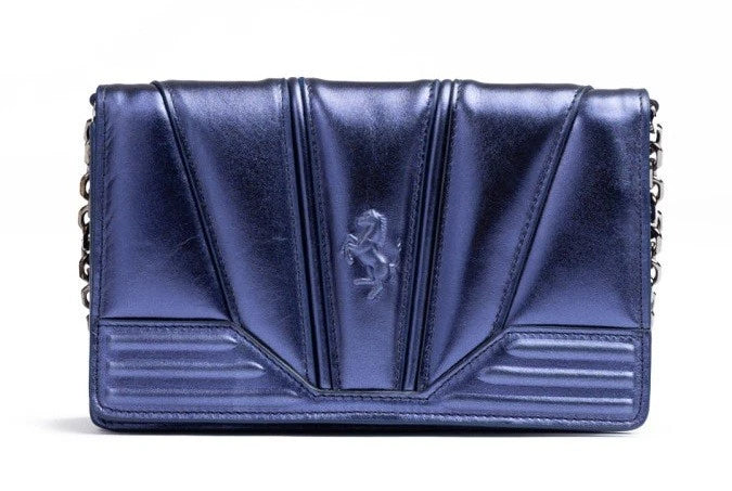 A metallic blue leather clutch bag with a central embossed Ferrari prancing horse logo, featuring vertical pleated detailing and a silver chain strap on the sides, set against a plain white background.